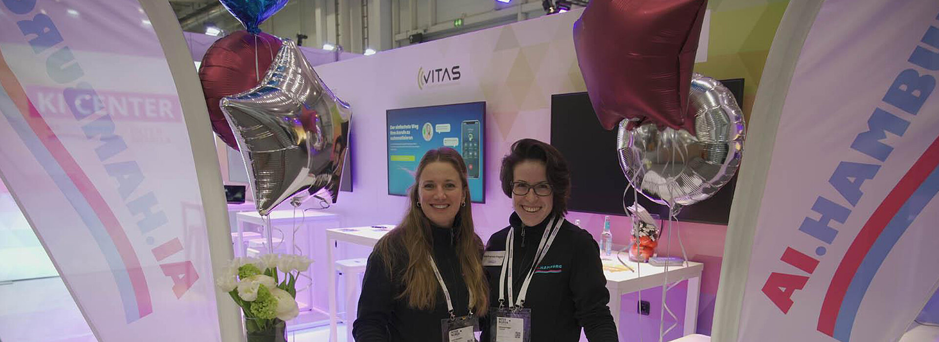 Blick auf Messestand des AI CENTERS. Zwei Personen im Vordergrund lächeln in die Kamera.