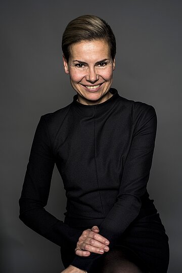 Porträtfoto von Anne Spielberg in schwarzer Kleidung vor neutralem, dunklem Hintergrund.