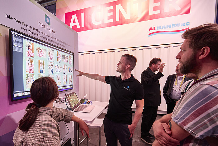 PHOTOPIA 2023 – Fair impressions AI CENTER A man is demonstrating the "Neurapix" software on a screen, which uses artificial intelligence to optimize photo editing processes, while several people are attentively watching.
