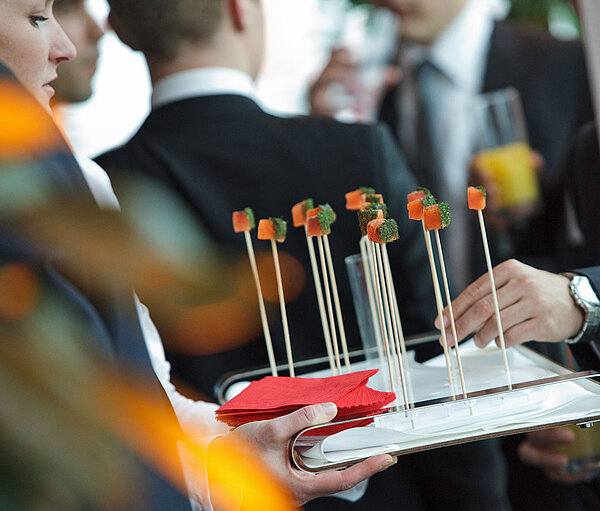Close-up of a tray with long sticks, holding a small treat at the end. A crowd is visible all around.