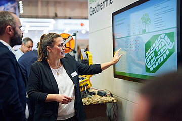 Eine Frau präsentiert auf einer Messe eine große digitale Anzeige zum Thema Geothermie. Sie zeigt auf ein Diagramm, während mehrere Personen aufmerksam zuhören.