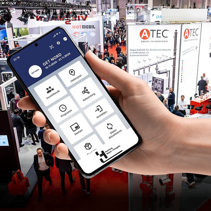 A hand holds a smartphone. The home screen of the GET NORD app can be seen on the screen with the areas “Exhibitors”, “Hall Plan”, “Program”, “Networking”, “Brands” and “Register”. In the background you can see a scene from the ongoing trade fair operations the GET NORTH.