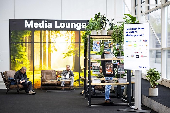 View from the center entrance to the Media Lounge. Several comfortable armchairs stand in front of a wall with a nature print and the words Media Lounge. Next to it are some shelves with magazines from media partners.