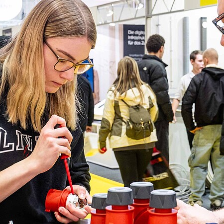 A young woman on the left side of the picture is concentrating and opening a small device with a wrench. A man on the right supports her.