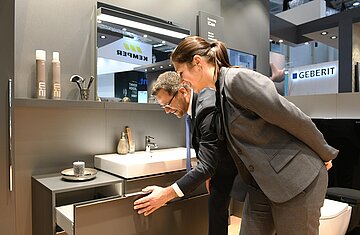 Ein Mann zeigt in einer Frau ein Vorführbadezimmer auf einer Messe. Er öffnet eine Schublade und beide betrachten diese.