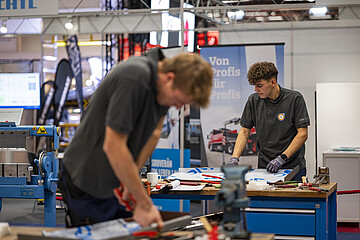 Two people are working at a workbench in a trade fair or exhibition hall. Tools and metal components are spread across the table. In the foreground, one person is shown out of focus while working on a metal piece, while another person is handling a workpiece at the bench. Machinery, exhibition stands, and a banner reading “Von Profis für Profis” can be seen in the background.