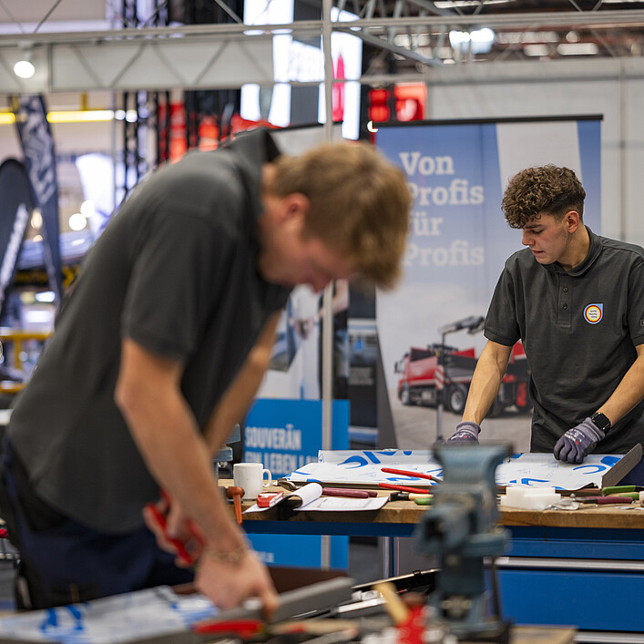 Two people are working at a workbench in a trade fair or exhibition hall. Tools and metal components are spread across the table. In the foreground, one person is shown out of focus while working on a metal piece, while another person is handling a workpiece at the bench. Machinery, exhibition stands, and a banner reading “Von Profis für Profis” can be seen in the background.